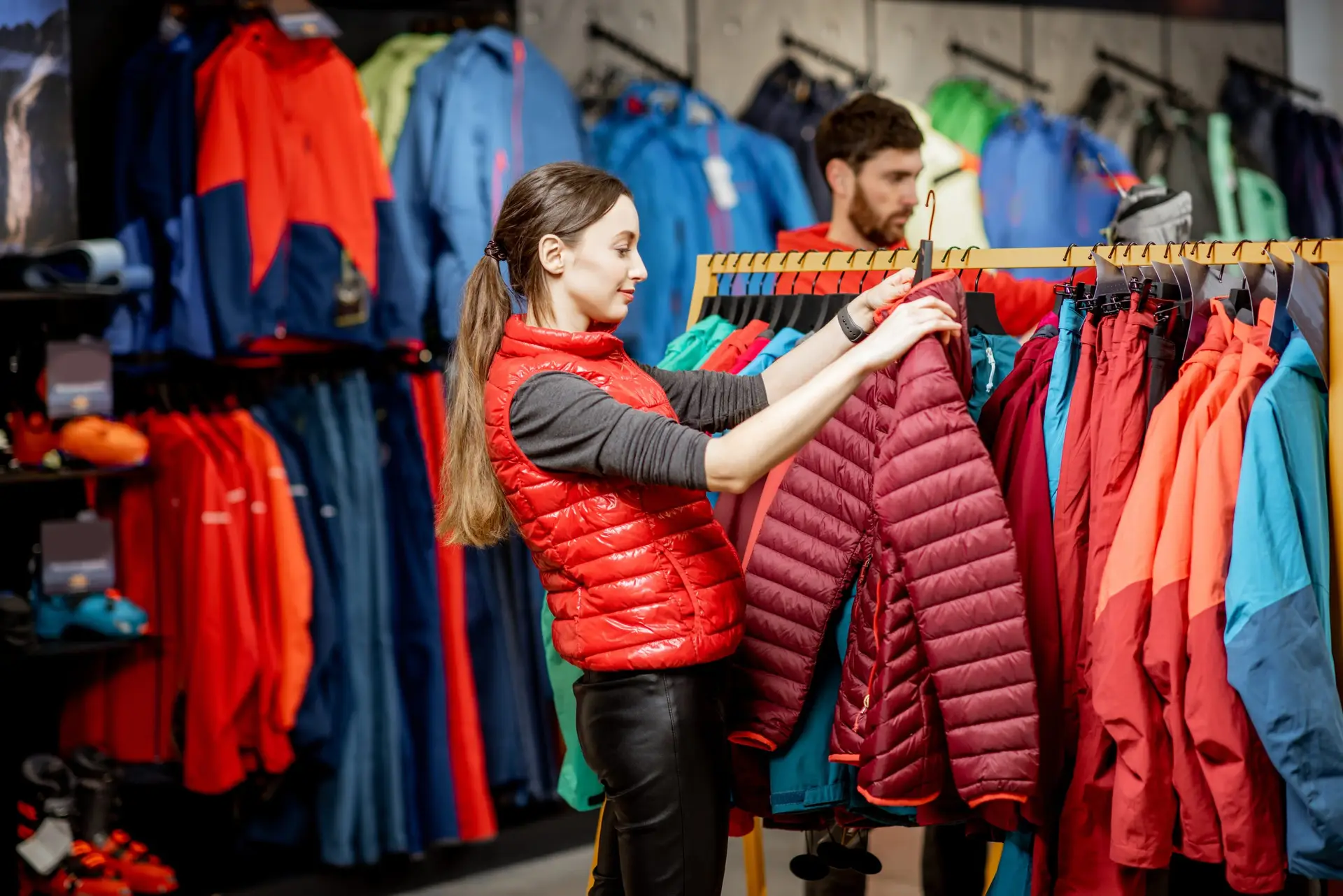 Young woman choosing winter clothes picking up down jackets on the hanger in the sports shop